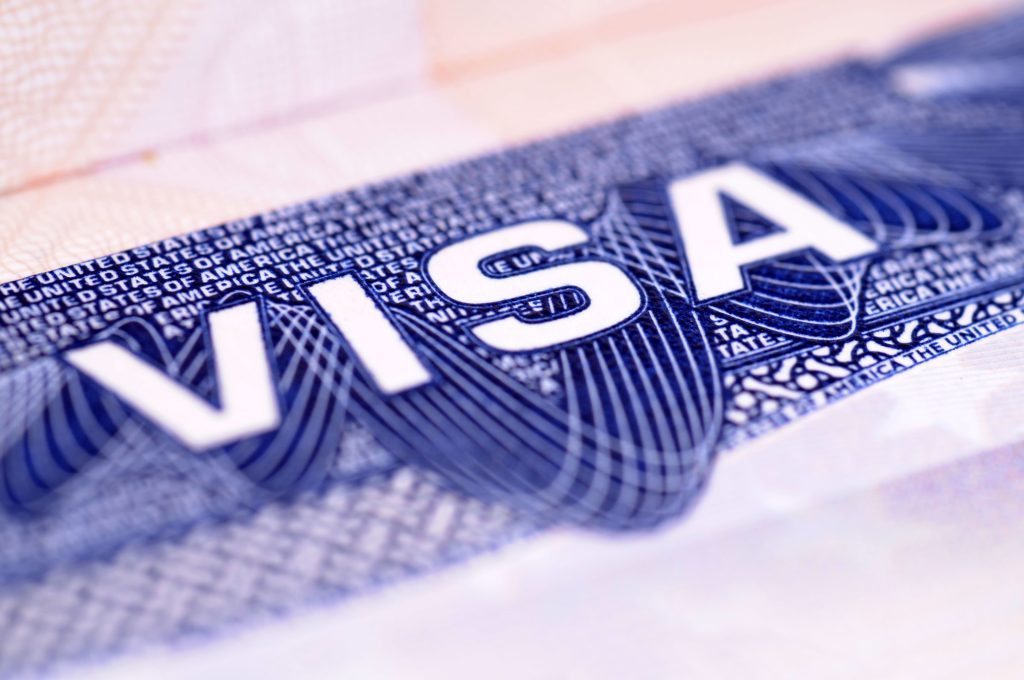 Close-up macro de um visto americano, com a palavra "VISA" em letras grandes brancas sobre fundo azul. Ao fundo, repetido em micro-impressão, lê-se "THE UNITED STATES OF AMERICA", junto a padrões guilhoché entrelaçados (linhas de segurança). O foco está nítido no centro e desfoca nas bordas.
